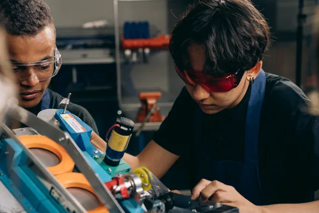 Two young engineers concentrating on precision work in a technical workshop, wearing safety goggles. Far approvare il contratto di apprendistato: obblighi e uffici cantonali
